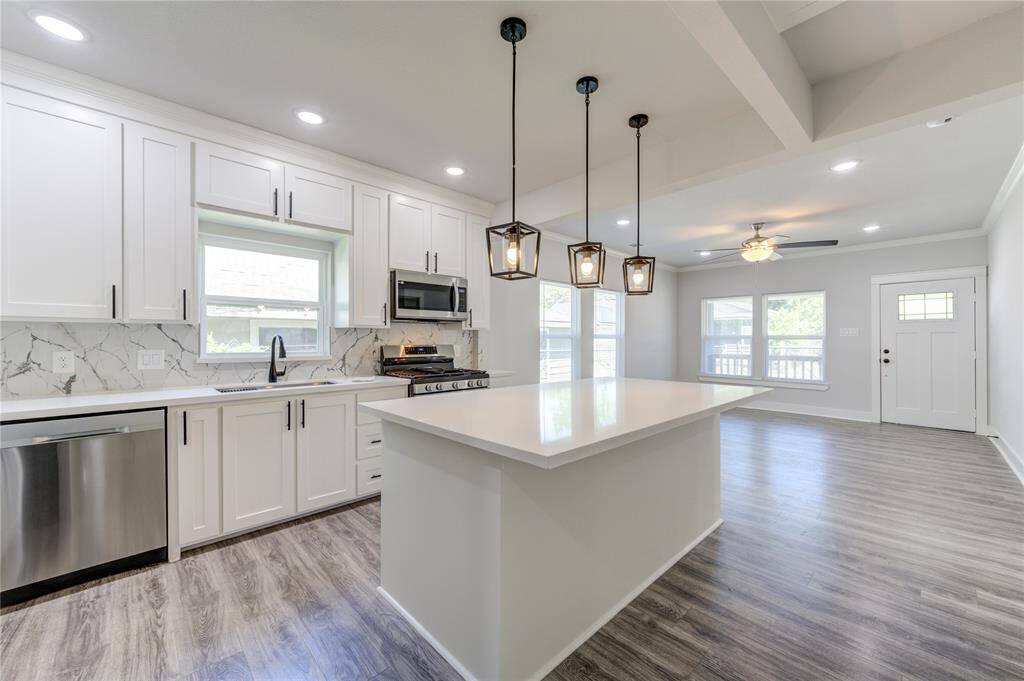 This modern kitchen features white cabinetry, stainless steel appliances, and a sleek marble backsplash. The open-concept layout includes a spacious island with pendant lighting, seamlessly connecting to a bright living area with large windows and a ceiling fan. The flooring is a contemporary wood finish, enhancing the overall elegance.