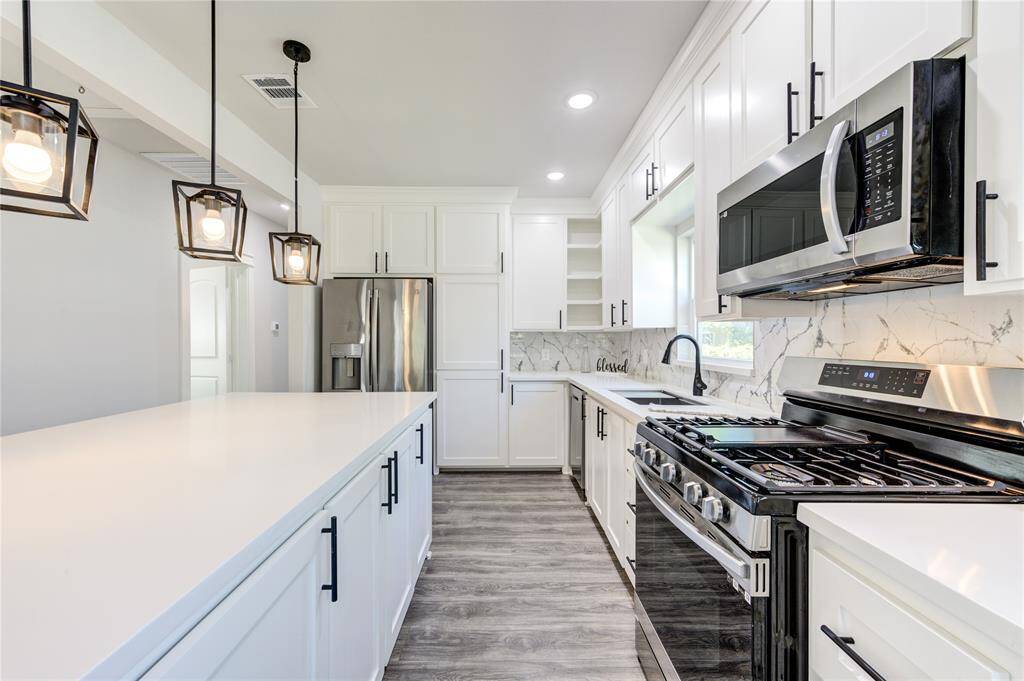 This modern kitchen features sleek white cabinetry, stainless steel appliances, and a spacious island with pendant lighting. The marbled backsplash and light wood flooring add elegance, while ample storage and counter space enhance functionality.