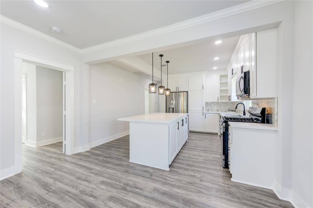 This modern kitchen features sleek white cabinetry, a spacious island with pendant lighting, and stainless steel appliances. The open layout and light wood flooring create a bright and inviting space.