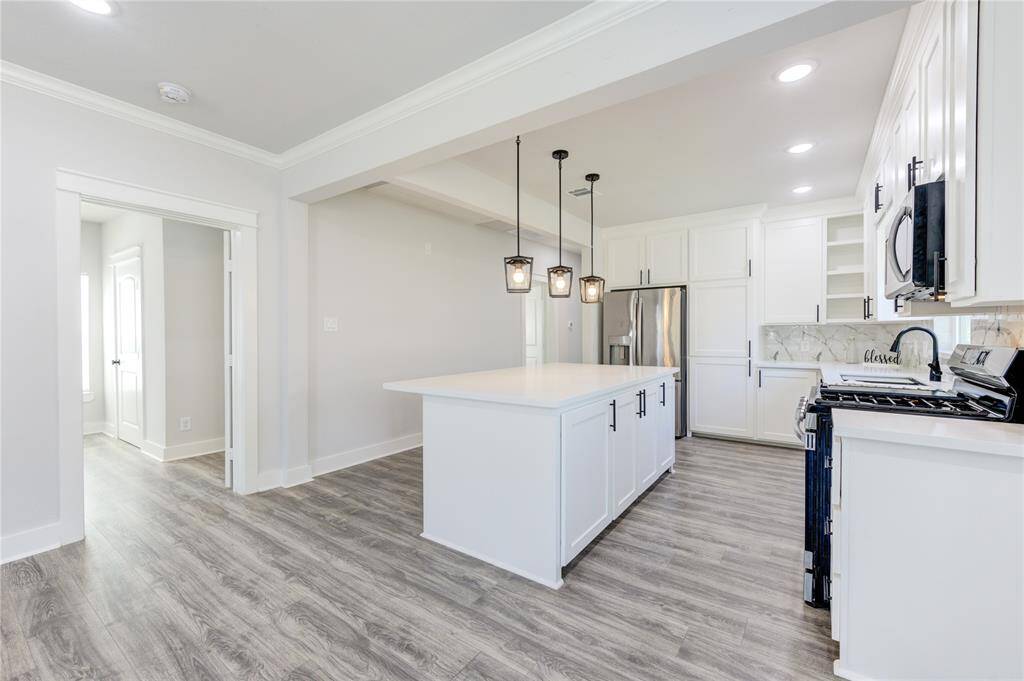 Bright, modern kitchen with white cabinetry, stainless steel appliances, and a large island. Features marble backsplash, pendant lighting, and wood-style flooring. Open layout connects seamlessly to adjacent rooms.