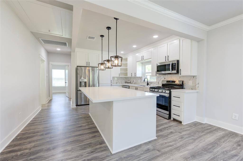 This modern kitchen features sleek white cabinetry, stainless steel appliances, and a spacious island with pendant lighting. The open layout and light wood flooring create a bright and inviting space, perfect for entertaining.