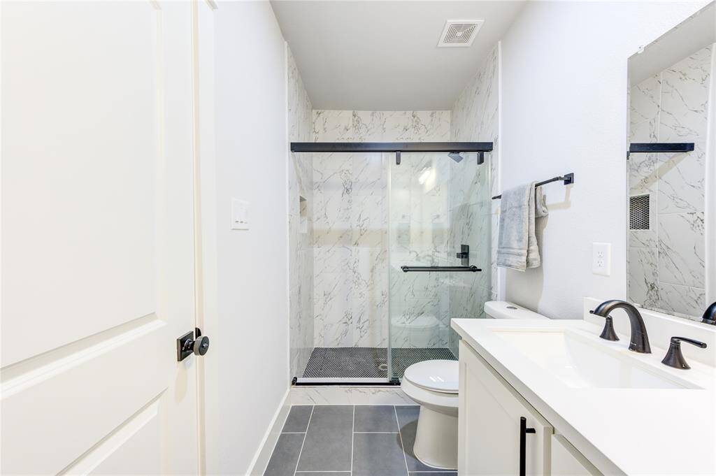 Modern bathroom with a sleek design, featuring a glass-enclosed shower with marble-patterned tiles. It includes a contemporary vanity with a white countertop and black fixtures, complemented by a large mirror and subtle lighting.