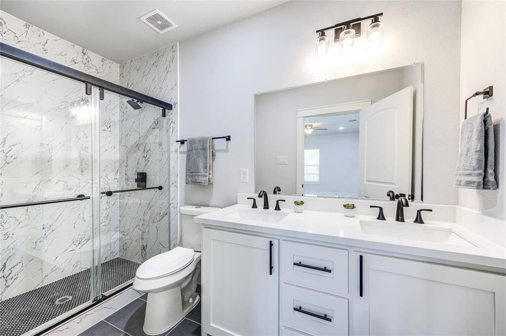This modern bathroom features a double vanity with sleek black fixtures, a large mirror, and ample storage. The spacious shower has a glass door and elegant marble-style tiling. The room is well-lit with contemporary lighting and accented with neutral tones.