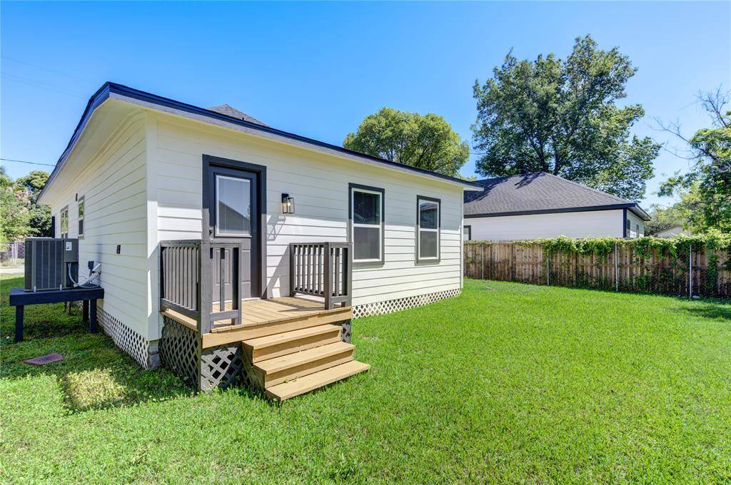 Charming backyard view of a home with a small deck and steps leading to a grassy yard. The house features white siding and black trim. A wooden fence and mature trees provide privacy.