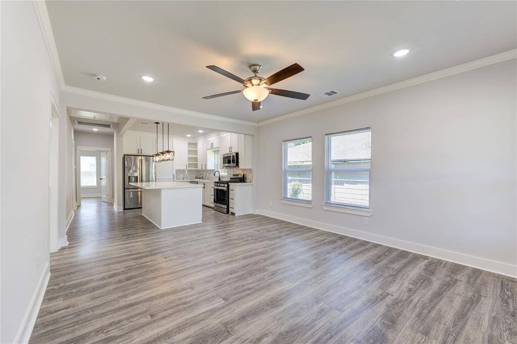 This photo shows a bright, open-concept living area with modern laminate wood flooring, recessed lighting, and a ceiling fan. The adjacent kitchen features stainless steel appliances, white cabinetry, and a central island with pendant lighting, providing a modern and welcoming space.