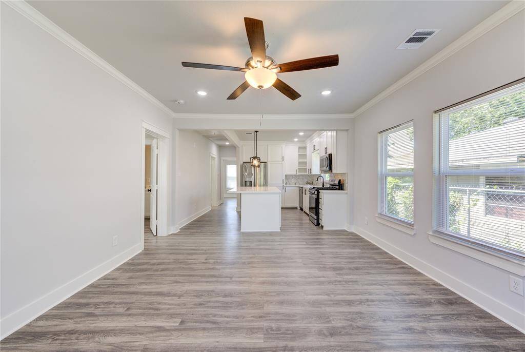 This photo showcases a bright, open-concept living area with modern laminate flooring, a ceiling fan, and large windows allowing natural light. The space extends into a sleek kitchen with white cabinetry, stainless steel appliances, and recessed lighting, creating a welcoming and contemporary atmosphere.