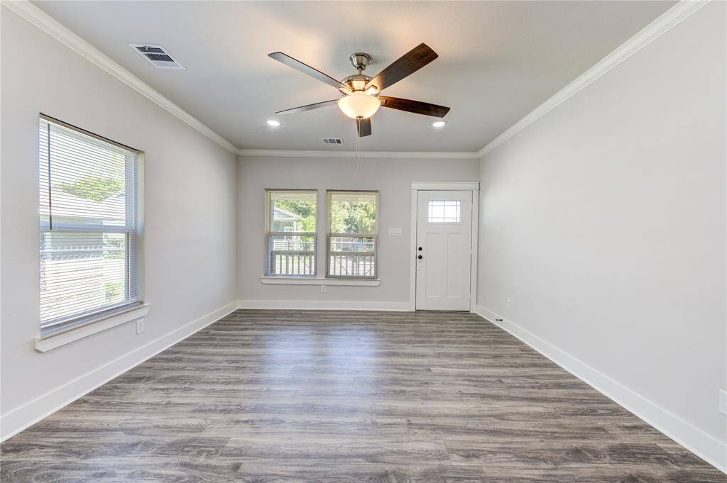 This photo shows a bright, open living room with light gray walls, wood-look flooring, and a ceiling fan. It features large windows for natural light and a front door with a window insert, creating a welcoming atmosphere.