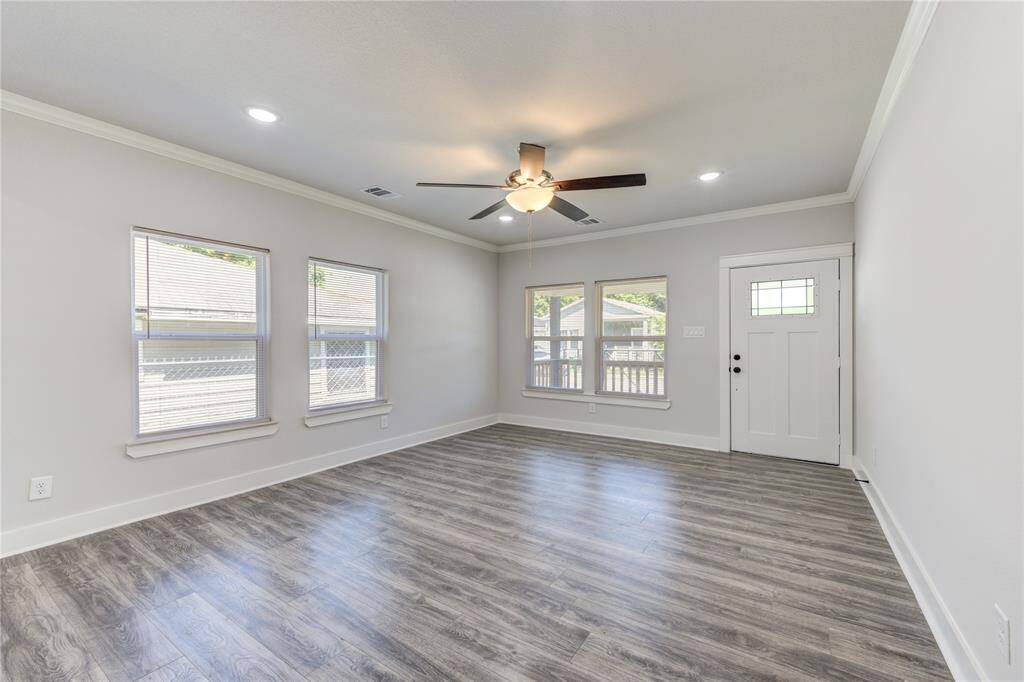 This bright, open living space features modern wood-look flooring, neutral walls, and recessed lighting. It includes a ceiling fan for comfort and multiple windows offering natural light. The room leads to a stylish entry door, creating a welcoming atmosphere.