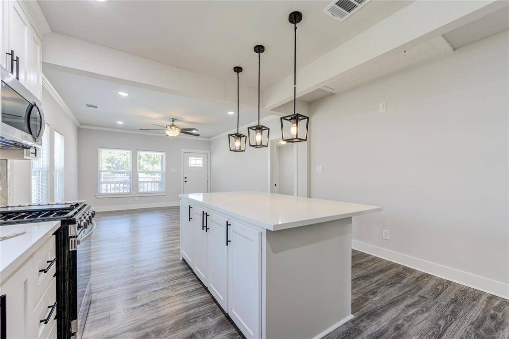 Modern kitchen with an open layout, featuring a large island with sleek white countertops. The space is bright with ample natural light from large windows. Stainless steel appliances and pendant lighting add a contemporary touch, while wood-look flooring extends into the living area.