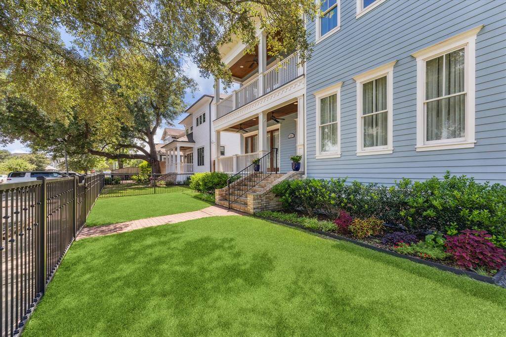 Serene Outdoor Escape in Houston Heights: A sprawling, perfectly manicured lawn stretches beneath mature shade trees, complemented by lush landscaping and a secure wrought-iron fence. This inviting outdoor space seamlessly blends natural tranquility with timeless architectural charm.