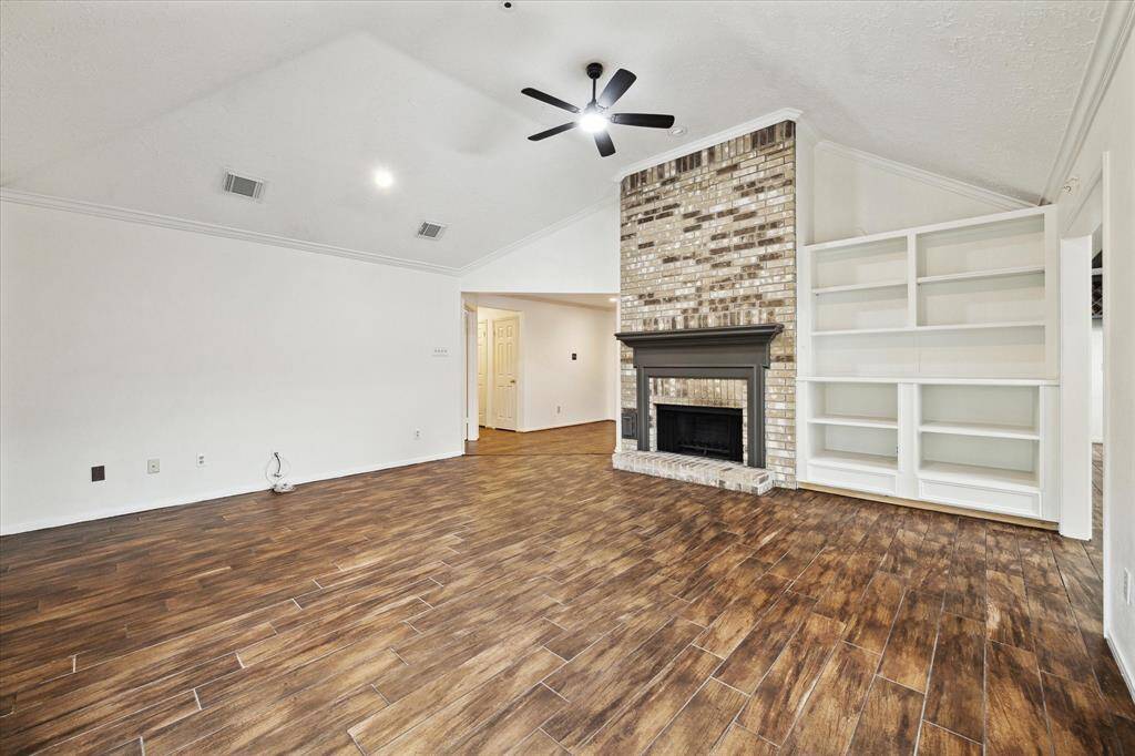The spacious living room features high, angled ceiling, wood-look tile flooring, a brick fireplace, and custom built-in shelves, offering a cozy yet modern vibe.