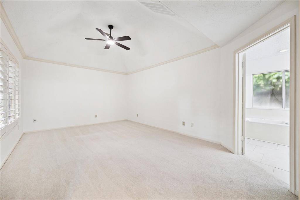 This spacious bedroom features high ceilings with a modern ceiling fan, neutral carpeting, and ample natural light from large windows with shutters. An open doorway leads to an adjoining bathroom, enhancing convenience and privacy.
