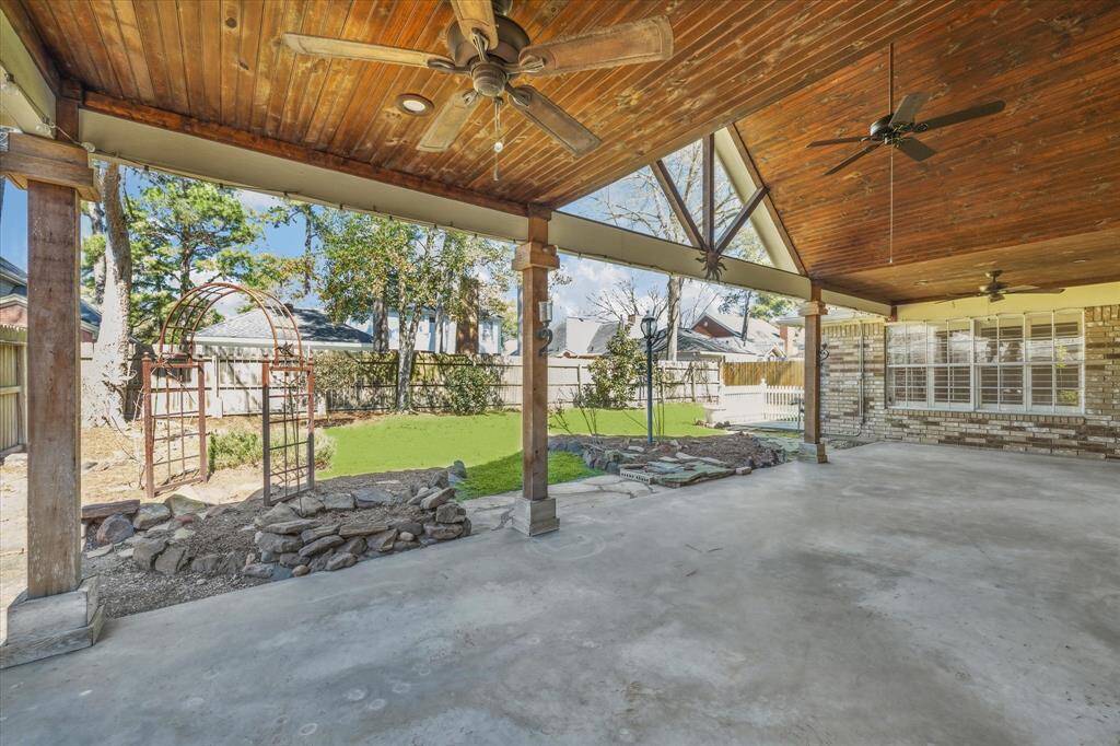 This dream covered patio is so spacious and features a wooden ceiling and multiple fans. It overlooks a well-maintained backyard with trees and a decorative arbor, perfect for outdoor relaxation and entertaining.