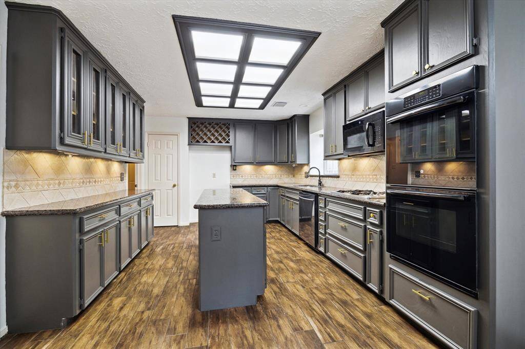 This kitchen features modern cabinetry with brushed gold hardware, sleek granite countertops, and a central island. It includes ample storage and under-cabinet lighting, all set on warm wood look tile flooring.