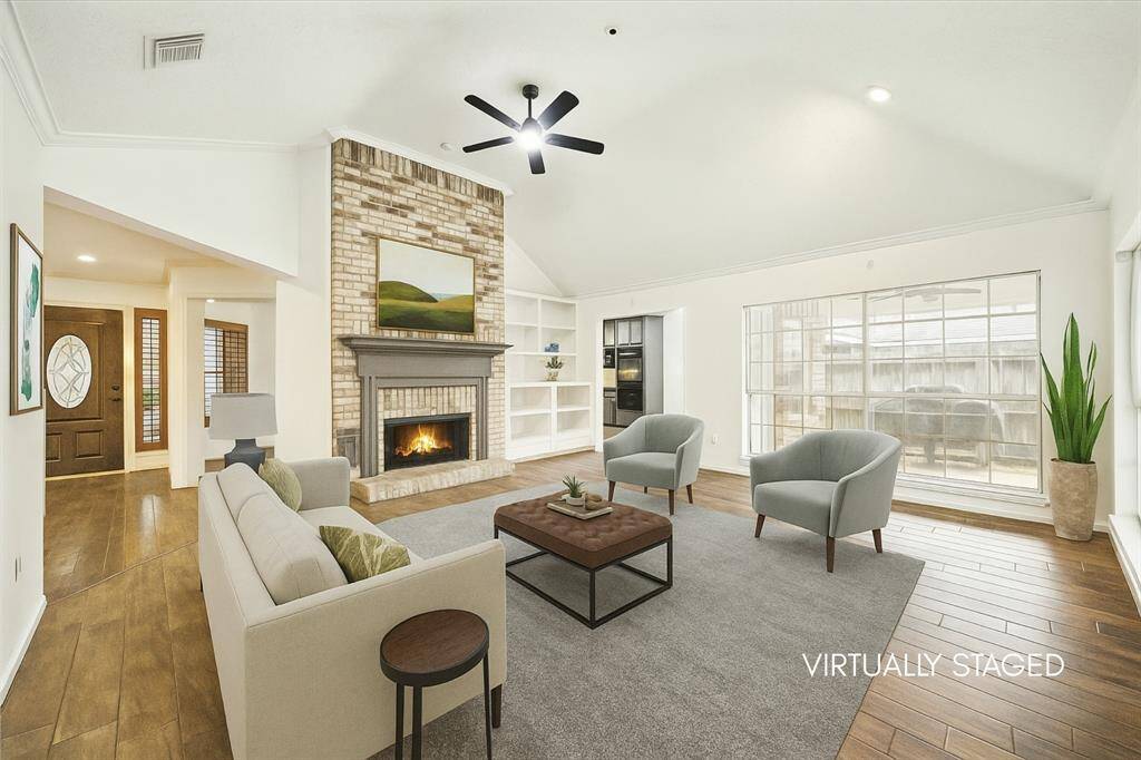 This virtually staged living room features a vaulted ceiling, a cozy brick fireplace, custom built-ins, and ample natural light from large windows. The open layout includes wood-look tile floors, and a welcoming entryway.