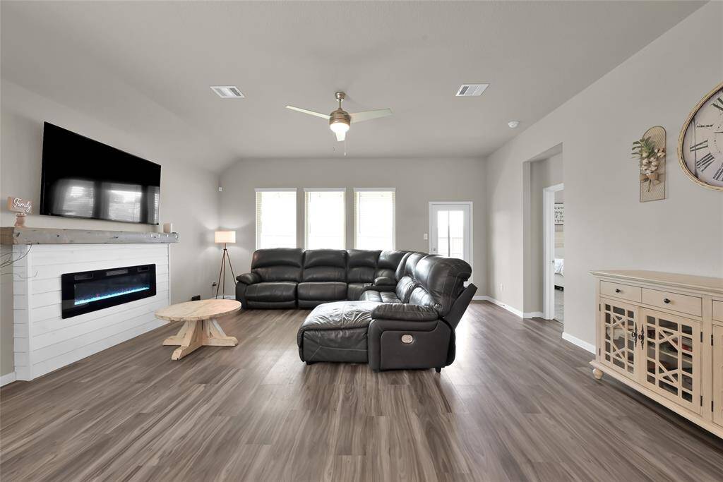 This is a spacious and modern living room with a sleek electric fireplace, large mounted TV, durable wood-look flooring, and comfortable leather seating. Bright and airy with ample natural light from the windows, the room features neutral colors, offering a versatile backdrop for personalization.