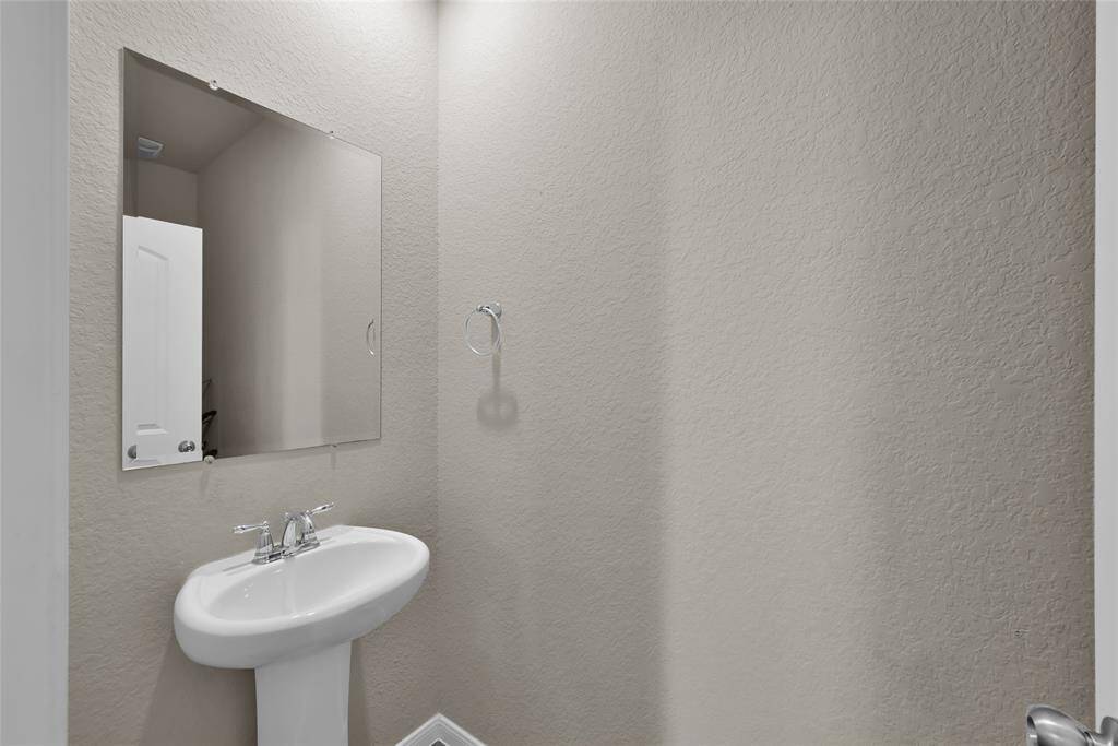 The photo shows a clean and simple half-bath with a pedestal sink, a large mirror, and a towel ring on a textured wall, presenting a compact and functional space.
