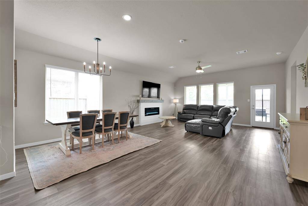 This is a spacious, open-plan living and dining area with modern lighting, large windows for natural light, wood-look flooring, and contemporary neutral decor. The space includes a sizable dining table set and a comfortable seating area with a sectional sofa, and there's direct access to the covered patio through back door.