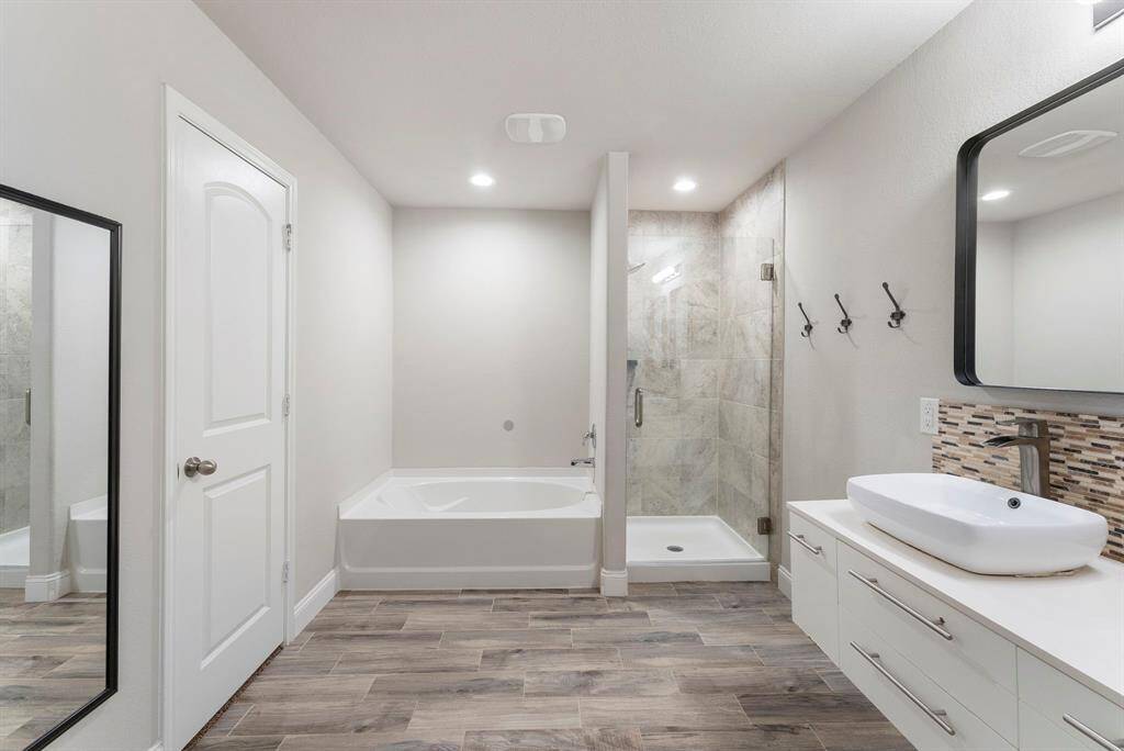 This modern bathroom features a spacious design with a separate shower and bathtub, stylish wood-look tile flooring, and a sleek white vanity with a vessel sink and contemporary mirror.