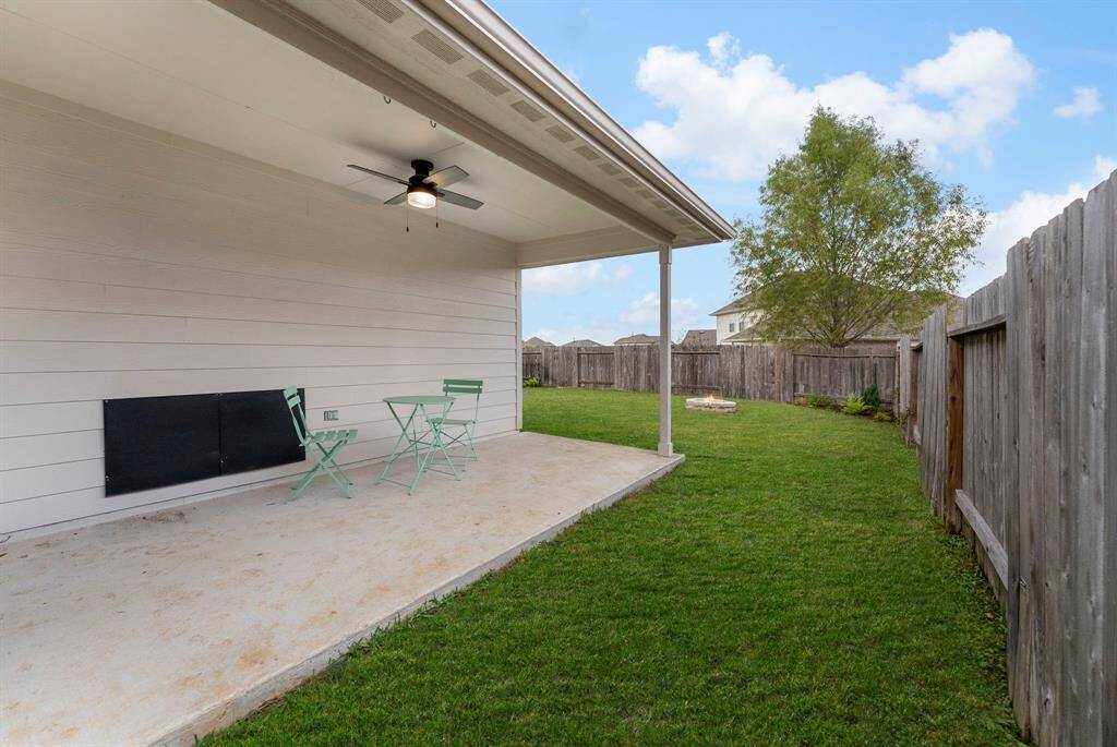 A cozy backyard with a covered patio, featuring a ceiling fan and seating area. The space is enclosed by a wooden fence, providing privacy.