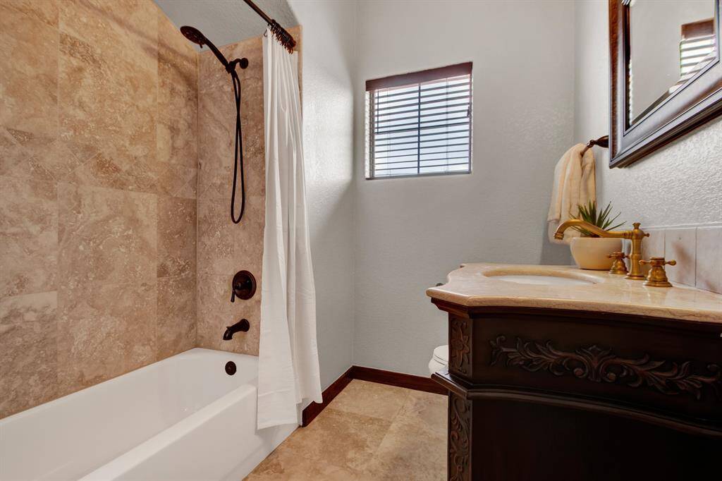 This full bathroom features a warm travertine tile surround, a bath tub, and a rich wood vanity with elegant brass and  bronze fixtures.