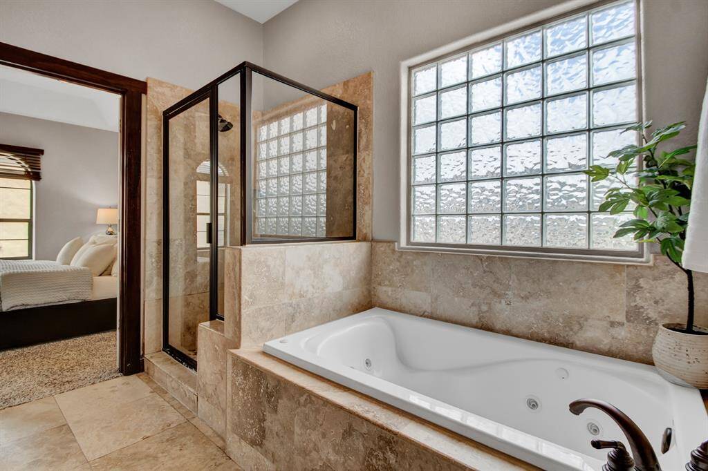 The spa-inspired primary bathroom features rich travertine tile, a jetted soaking tub beneath a large privacy glass window, and a walk-in shower just steps away for a truly luxurious retreat.