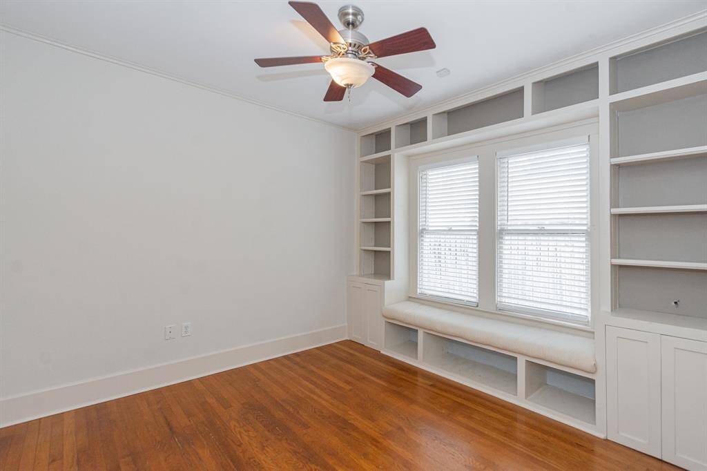 Cozy secondary bedroom with hardwood floors, built-in shelving, and a window seat. Features a ceiling fan and ample natural light through large windows. Perfect for a reading nook or small home office.