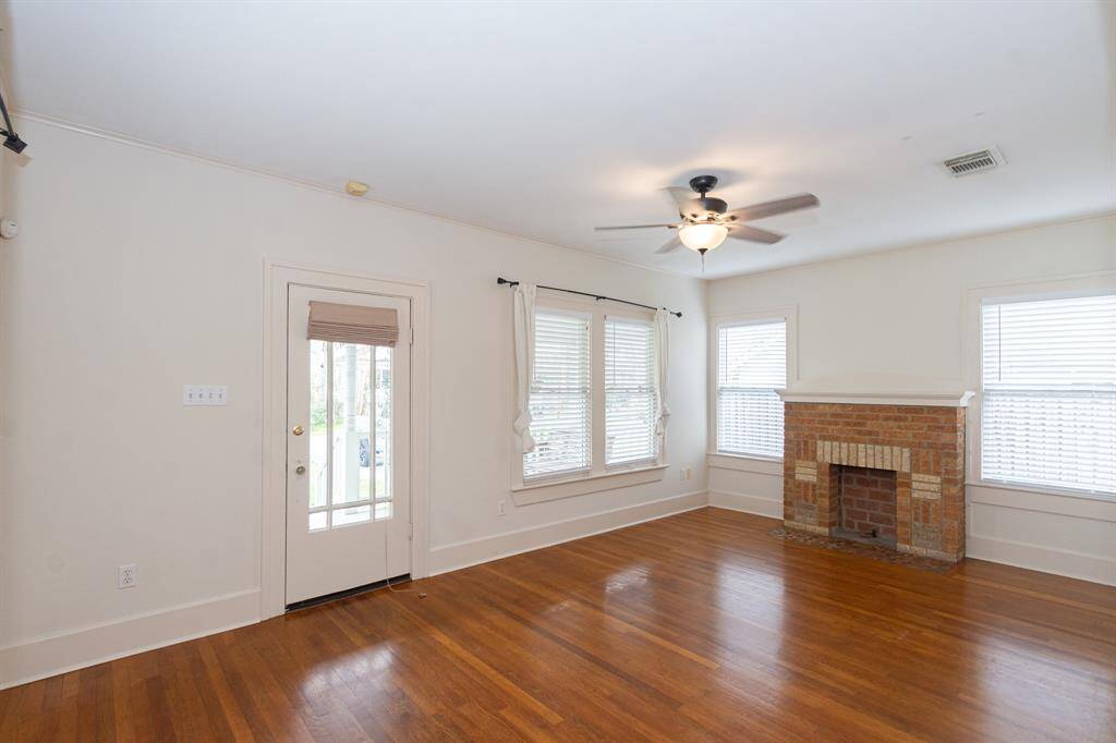This spacious living room features rich hardwood floors, abundant natural light, and a charming brick faux fireplace as the focal point.