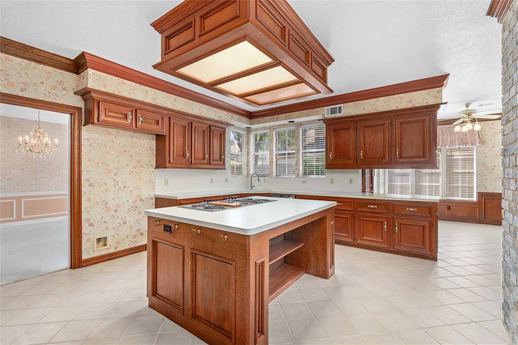 Your large kitchen has rich wood cabinetry with matching crown molding, a central island with a cooktop, and ample natural light from large windows. The adjoining breakfast nook is visible giving this classic design an inviting feel.
