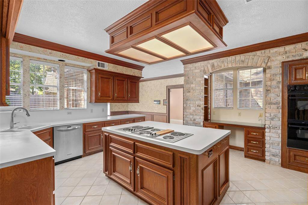 Your new spacious kitchen features rich wooden cabinetry with matching crown molding, a central island with built-in cooktop, and ample counter and storage space. The room is well-lit with natural light from multiple windows and is complemented by a brick accent wall and tile flooring.