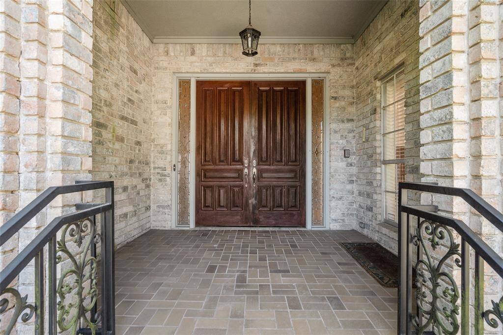 Your grand entrance has elegant double wooden doors flanked by decorative sidelights. The porch features brick walls, a tiled floor, and ornate wrought iron railings creating a welcoming and sophisticated entryway.