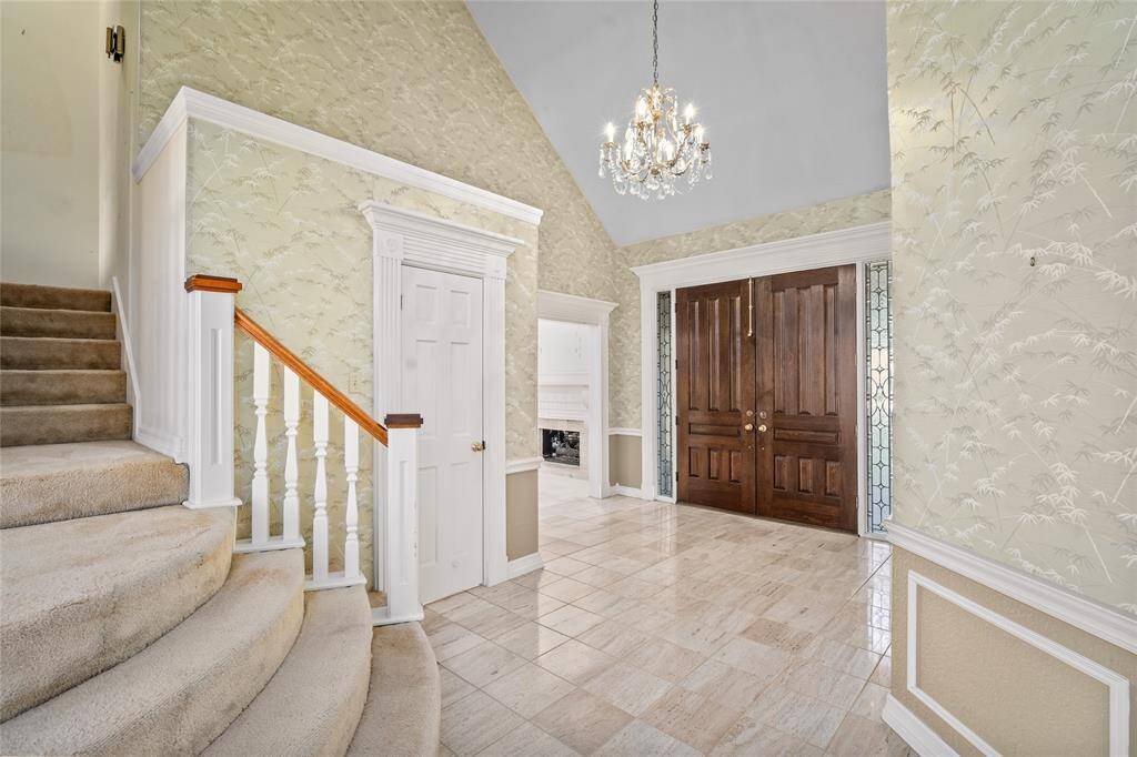 Your classy foyer features a high ceiling, a grand chandelier, decorative wainscoting and tile floors. The space is highlighted by the striking wooden double door entry and a carpeted staircase with a classic wooden railing, creating a warm and cultured entrance.