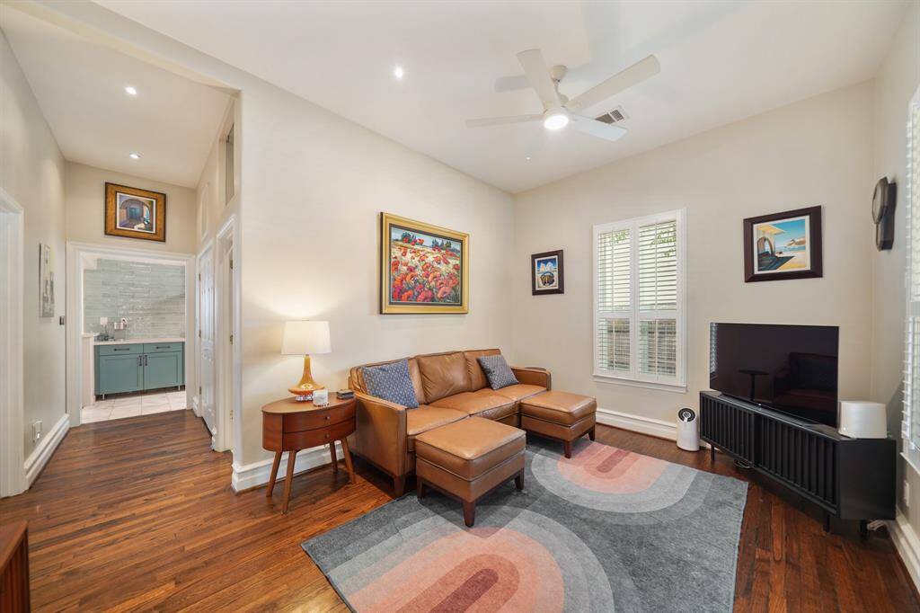 This view of the living room features warm hardwood floors, a modern ceiling fan, and has ample natural light from the window. The adjacent hallway leads to a stylish bathroom.