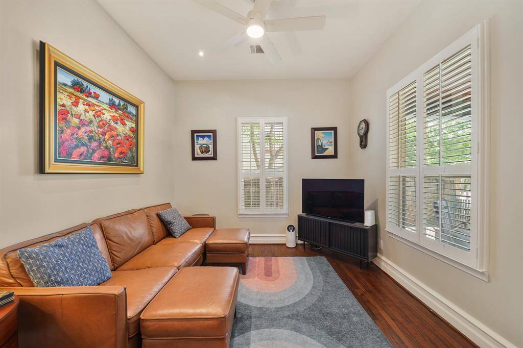 Cozy living room with a warm, inviting atmosphere. Large windows with shutters allow natural light, highlighting the hardwood floors. Perfect for relaxation and entertainment.