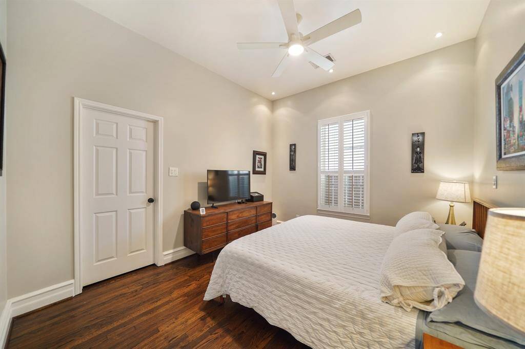 This bedroom features warm hardwood floors, a cozy bed, and a ceiling fan. It includes a window with shutters for natural light, creating a serene and inviting atmosphere.