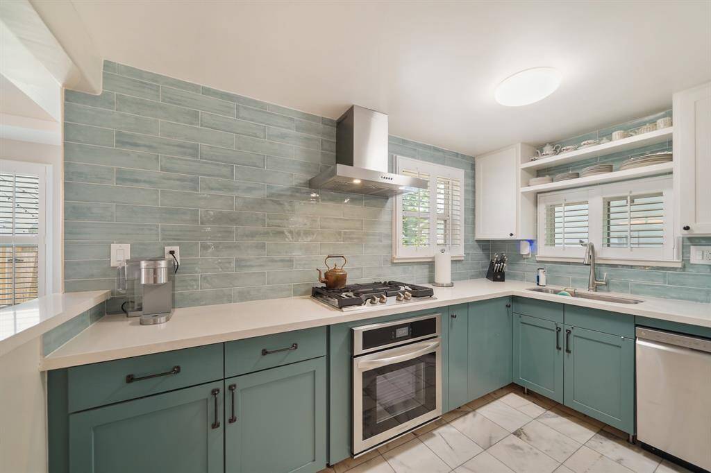 This kitchen features a modern, clean design with teal cabinets and a light blue tile backsplash. It includes stainless steel appliances, a gas stove, and ample counter space. The room is brightened by natural light from two windows with shutters, and open shelving provides additional storage.