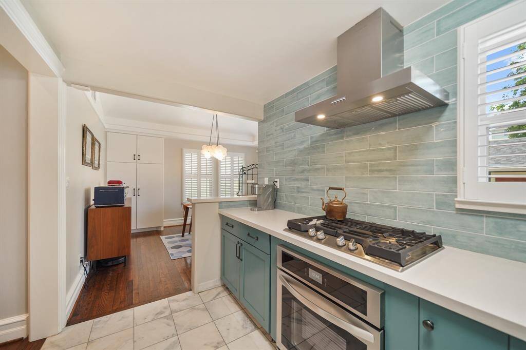 This photo showcases a modern kitchen with teal cabinetry, a sleek gas stove, and a stylish hood. It features a light blue tile backsplash, marble floors, and a bright dining area with hardwood flooring and a pendant light. The space is airy and well-lit, offering a blend of functionality and design.