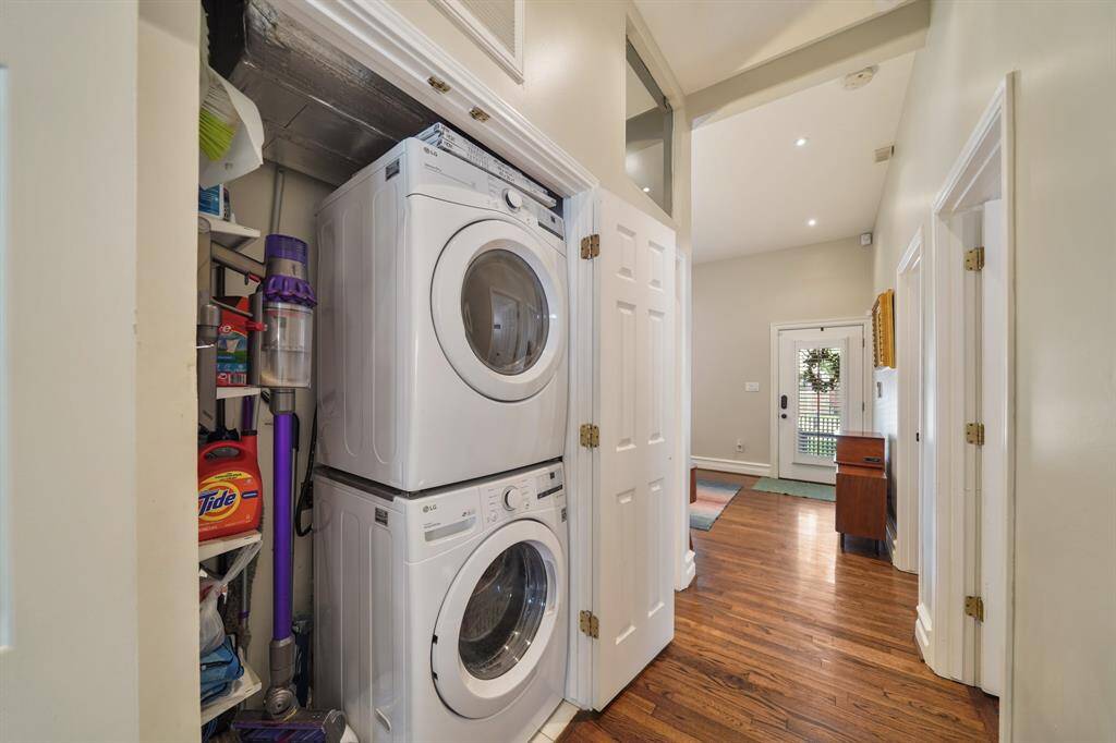 This photo showcases a compact laundry area with a stacked washer and dryer in a hallway closet. The space is efficiently utilized with additional shelving for storage. The hallway features hardwood flooring and leads to a bright entryway with a glass door, enhancing the home's welcoming atmosphere.