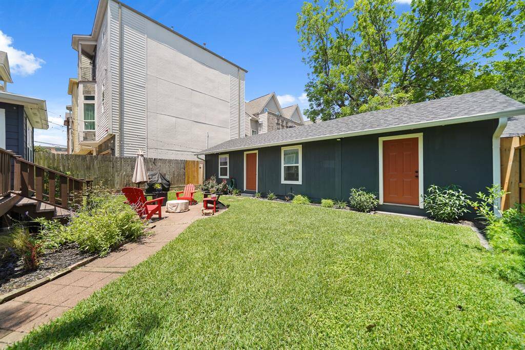 This backyard features a well-maintained lawn with a paved pathway leading to a cozy seating area with red Adirondack chairs and a fire pit and features a guest house with double entry access, surrounded by small shrubs and trees, providing a private and inviting outdoor retreat.