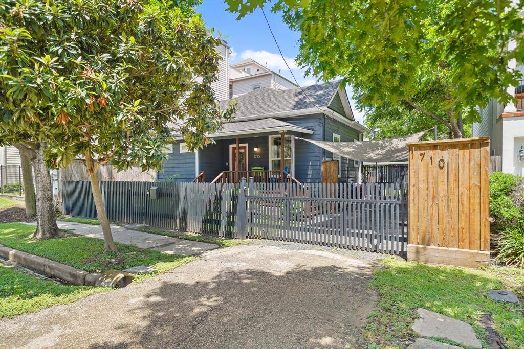 Charming single-story home with a dark exterior, nestled among lush trees. It features a welcoming front porch and a gated wooden fence for added privacy, creating a cozy and inviting atmosphere.