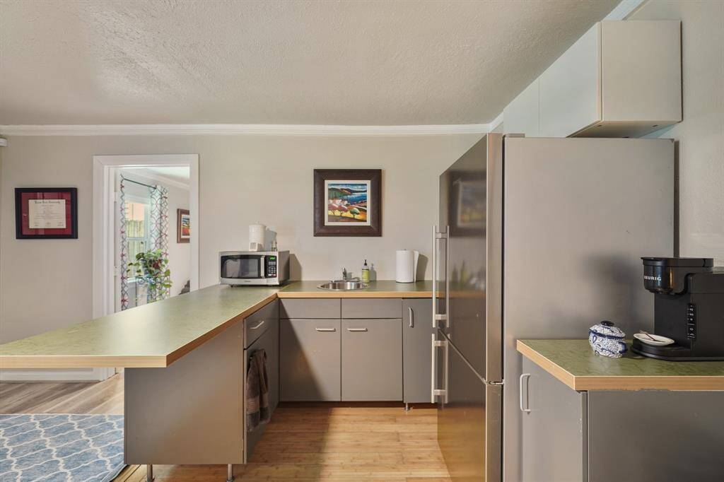 Cozy kitchen with modern gray cabinetry, a spacious countertop, and stainless steel appliances. Features a small sink, microwave, and coffee maker. Natural light brightens the space, with an adjacent room visible through a doorway.