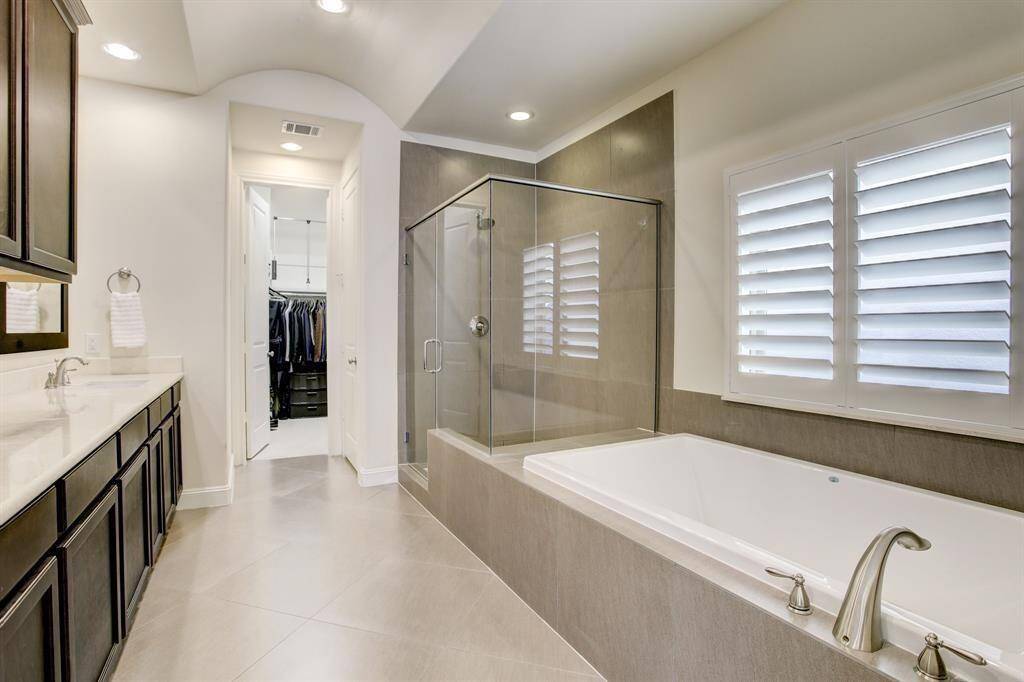 This is a spacious, modern bathroom featuring a glass-enclosed shower and a deep soaking tub. It includes dual sinks with dark cabinetry and has ample natural light from the shuttered window. The room leads into a walk-in closet.