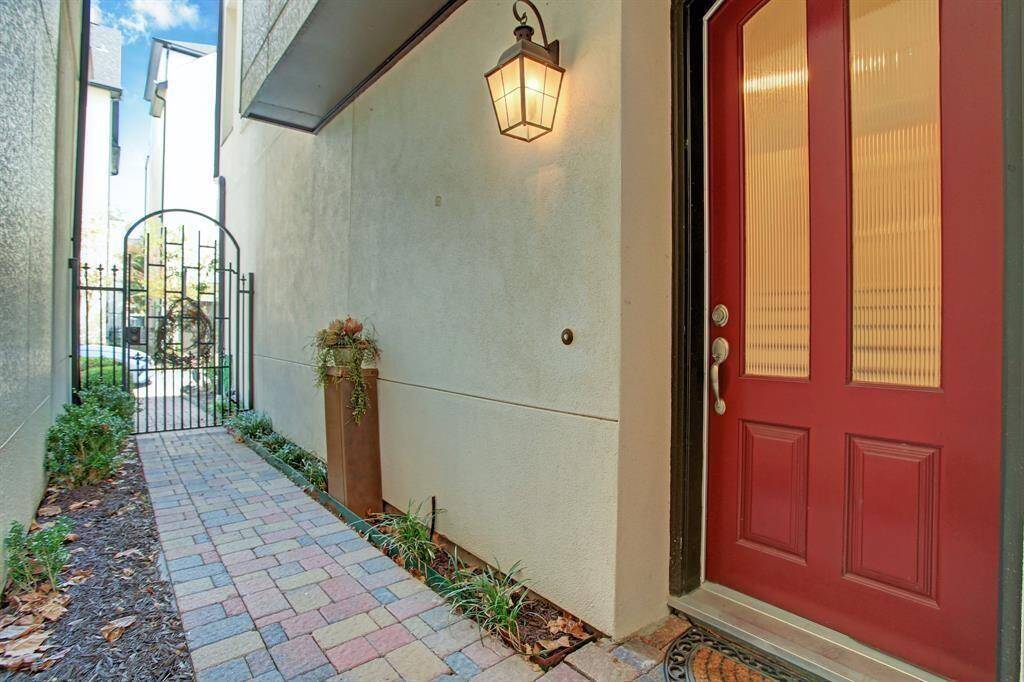 Inviting entry with a red door and decorative glass panels, leading to a narrow walkway lined with plants. The pathway is paved with multi-color bricks and features a wrought iron gate, creating a welcoming entryway.
