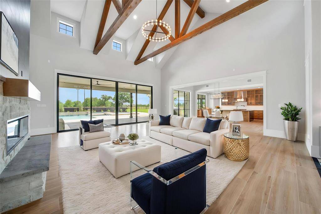 A grand welcome into the living area featuring high vaulted ceilings with exposed wooden beams, a modern electric fireplace, and large sliding glass doors that open to a patio and pool area. The open floor plan seamlessly connects to a contemporary kitchen, providing a bright and airy ambiance.