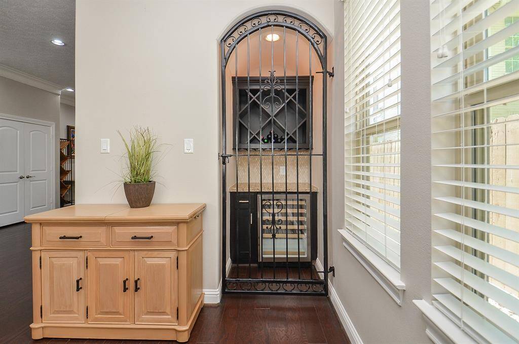This photo features a stylish wine storage area with an ornate iron gate, wood cabinetry, and a small countertop. It's located next to a large window with blinds, providing natural light and a cozy atmosphere.