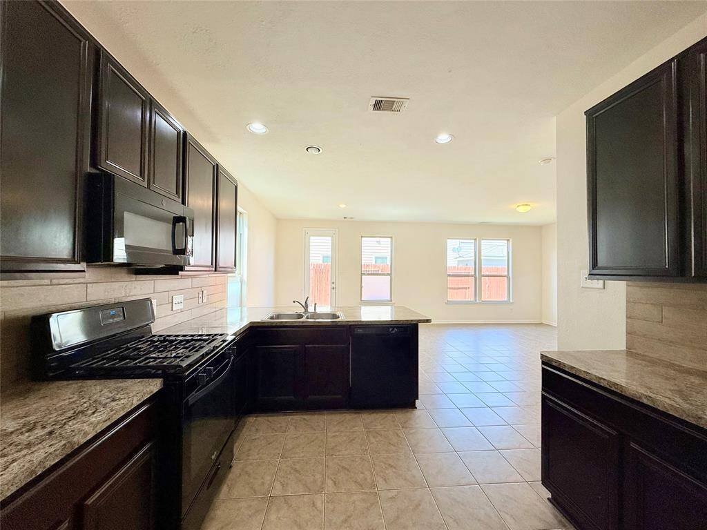 This photo showcases a bright kitchen with dark cabinetry, granite countertops, and stainless steel appliances. It opens to a spacious, tiled living area with large windows and a door leading to the backyard.