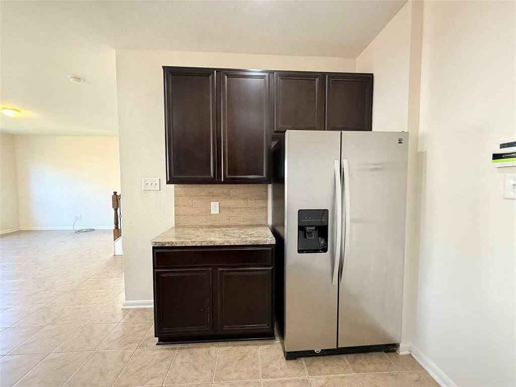 This photo shows a kitchen area with dark wood cabinets, a stainless steel refrigerator, and beige tiled flooring. The space is bright and opens into an adjoining room.