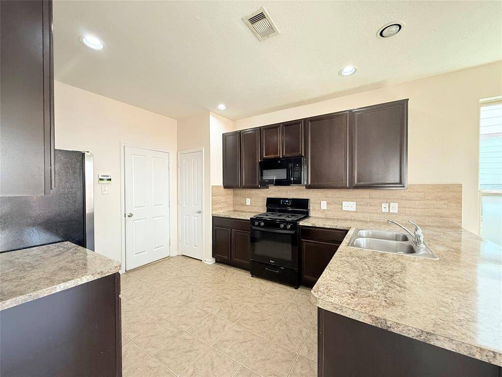 Modern kitchen with dark wood cabinets, granite countertops, and stainless steel appliances. Features a gas stove, built-in microwave, and dual sink. Tile flooring and ample natural light enhance the space.