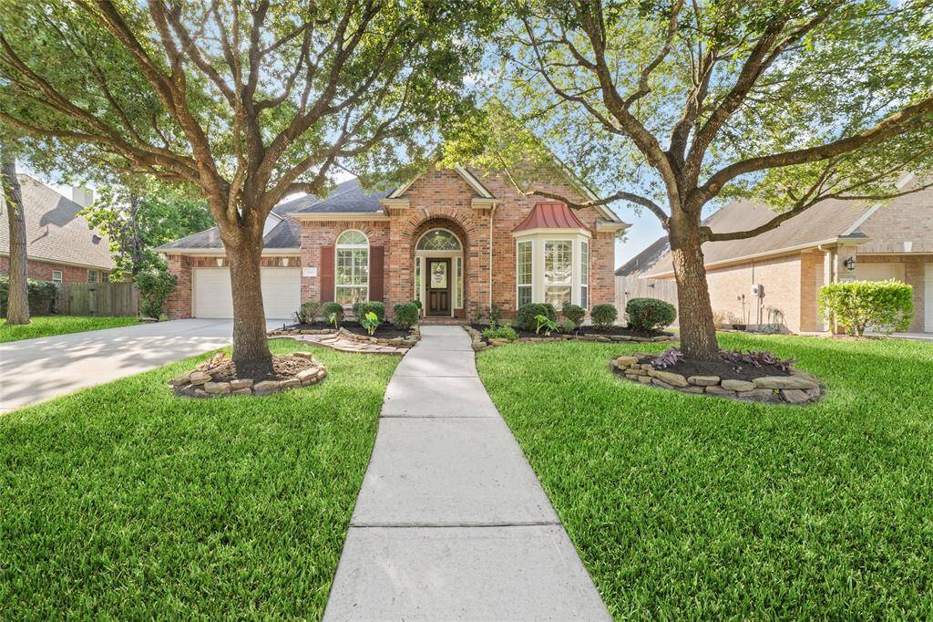 Charming brick home on all four sides with a welcoming arched entryway, nestled between mature trees. The well-maintained lawn and landscaped garden enhance its curb appeal. The driveway leads to a spacious three-car tandem garage.