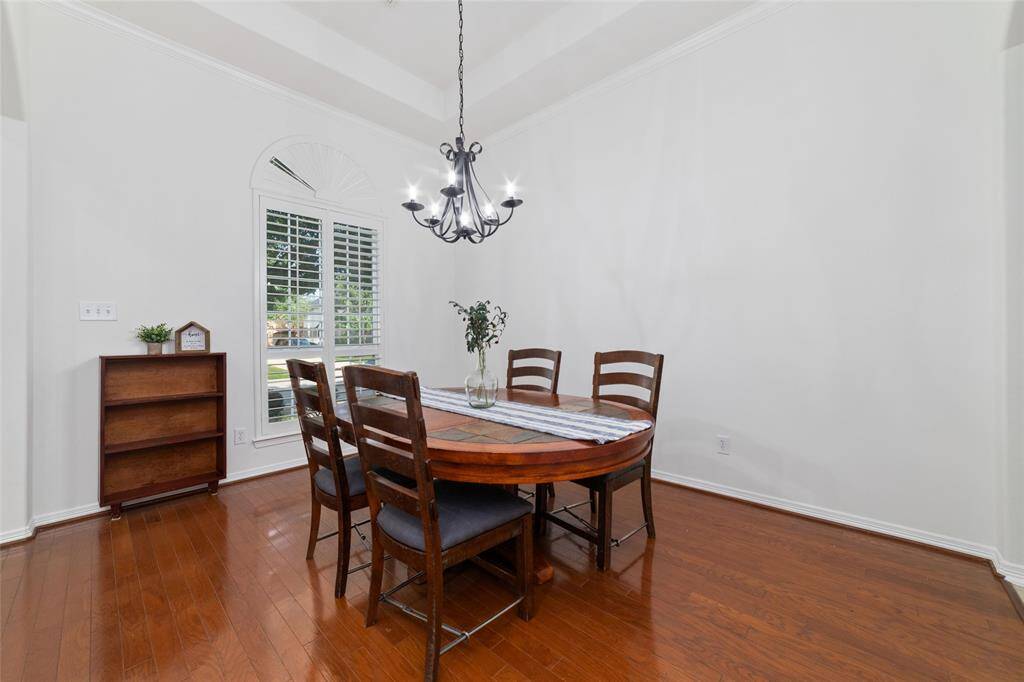 This dining room features a warm, inviting atmosphere with hardwood floors, a stylish chandelier, and a large window with plantation shutters for natural light. Plenty of space for a large dining table.